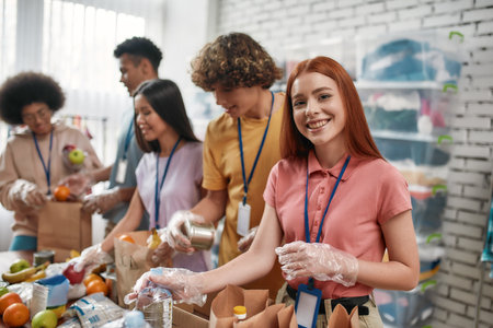 Young female volunteer smiling at camera while packing food and drinks donation into paper bags and box, Small group of people working in charitable foundationの写真素材