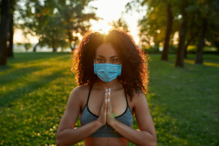 Stay calm. Young mixed race woman wearing protective face mask keeping palms together and looking at camera while practicing yoga outdoors in nature during covid19 pandemicの写真素材