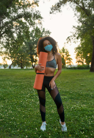 Time for yoga. Full leghth of a young mixed race fitness woman wearing protective face mask holding yoga mat and looking at camera while standing in park in the morningの写真素材