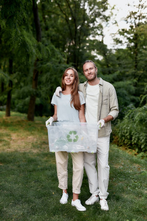 Vertical shot of young happy couple, eco volunteers holding recycle bin and looking at camera while collecting plastic waste together in the green forestの写真素材