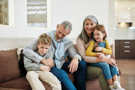 Grandparents with grandchildren hugging and smiling while sitting together on sofa in the living...