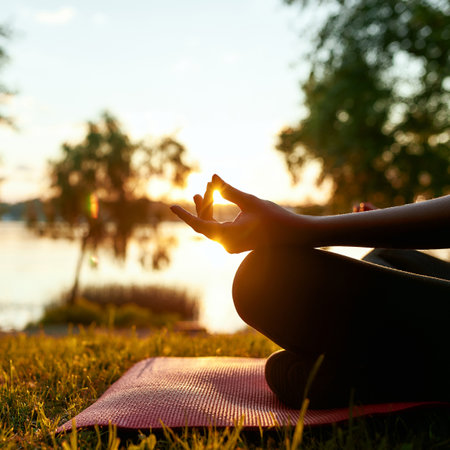 Cropped shot of a woman doing yoga outdoors near lake or river at sunrise in the morning, sitting in lotus pose on the background of nature and meditatingの写真素材