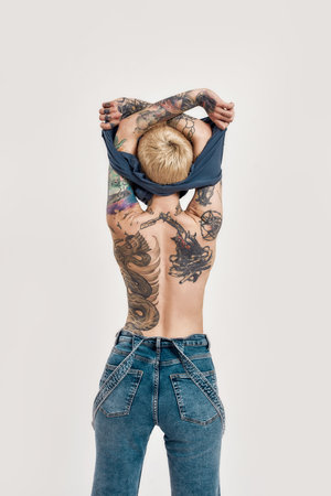 Tattoo and piercing. A tattooed and pierced white girl wearing denim overall standing with her back to a camera in a process of taking off a blue t-shirtの写真素材