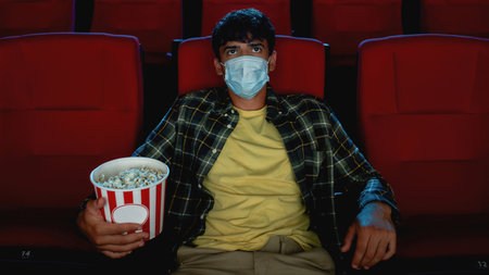 Young guy in casual clothes wearing safety mask, holding popcorn basket while watching movie, visiting cinema alone during coronavirus pandemicの写真素材
