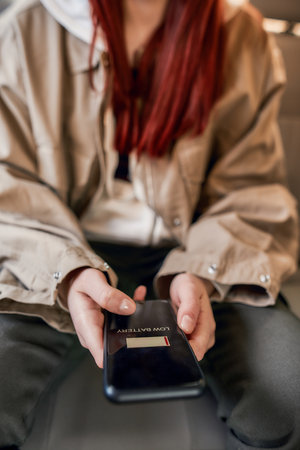 Close up of female hands holding mobile phone while sitting on back seat in the car. Low battery notification on the screenの写真素材