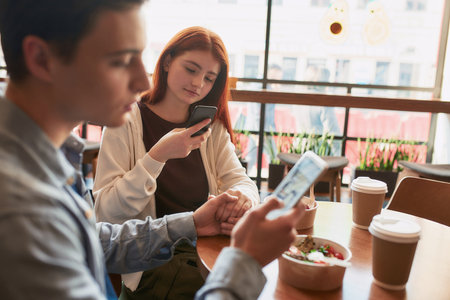Close up of teenage couple holding hands while girlfriend taking pictures with lunch, using smartphone, sitting in a cafe on a daytimeの写真素材