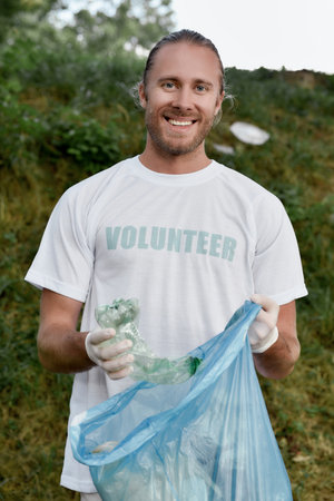Recycling concept. Vertical shot of a smiling young man, male volunteer wearing uniform and rubber gloves collecting plastic waste in the forest or parkの写真素材