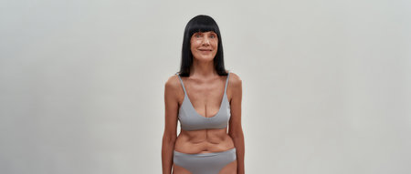 Attractive middle aged caucasian woman in lingerie smiling at camera while standing against grey background, panoramic bannerの写真素材