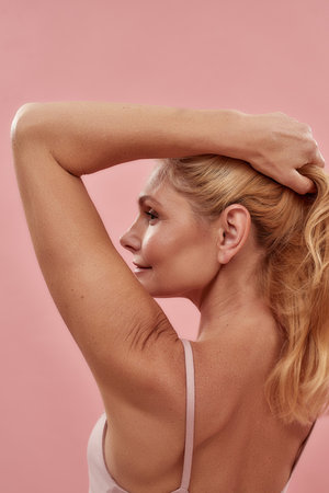 Natural beauty. Close up portrait of a beautiful half naked mature blonde woman holding hair and looking away while posing in studio over pink backgroundの写真素材