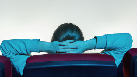 Back view of relaxed guy leaning on the back of chair and keeping his hands over the head while watching a movie alone in empty theater auditoriumの写真素材