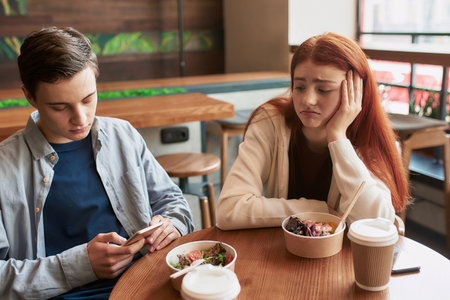 Two teenagers spending time together, sitting in a cafe on a daytime. Upset girl looking at her...
