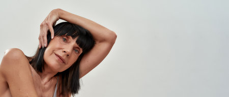 Aging process. Portrait of caucasian senior woman keeping one hand behind head, smiling at camera while posing against grey backgroundの写真素材