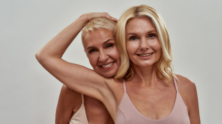 Studio portrait of two happy blonde women smiling at camera, standing together, posing isolated over gray backgroundの写真素材