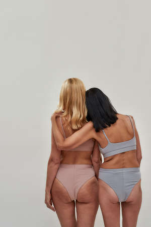 Back view of two caucasian women in underwear standing together while posing over light background. Warmth in relation, mature beauty conceptの写真素材