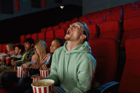 Young guy catching popcorn with his mouth while watching a movie in the cinema with friends