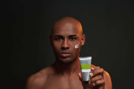 Portrait of shirtless african american man looking at camera, holding beauty product while posing with cream applied on his cheek isolated over black backgroundの写真素材