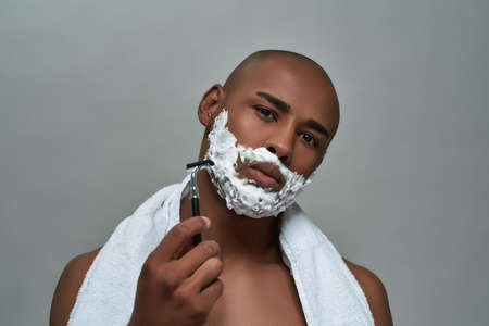 Handsome young african american man with shaving foam applied on his face looking at camera, holding steel razor, posing isolated over gray backgroundの写真素材