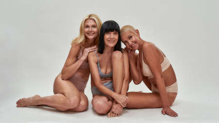 Three caucasian senior women in underwear smiling at camera while posing half naked in studio, sitting together isolated over light backgroundの写真素材