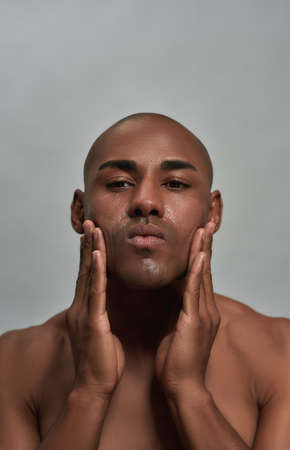 Portrait of shirtless young african american man looking at camera while applying cream on his face, posing isolated over gray backgroundの写真素材