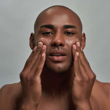 Closeup portrait of handsome young african american man looking at camera, applying cream on his face, posing isolated over gray backgroundの写真素材