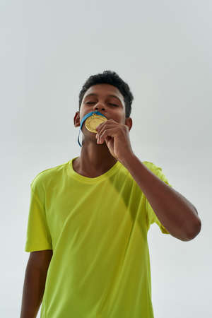 Vertical shot of a teenage african boy kissing gold medal with closed eyes while standing isolated...