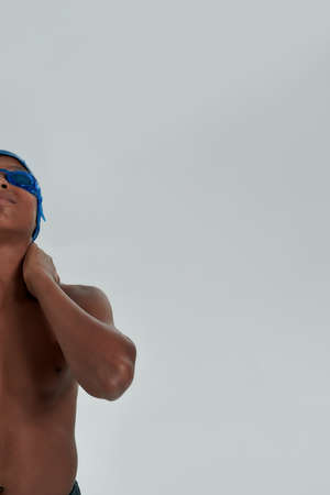 Half face portrait of a teenage african boy in swimming cap and goggles keeping arms on his neck and looking upside while posing isolated over grey backgroundの写真素材