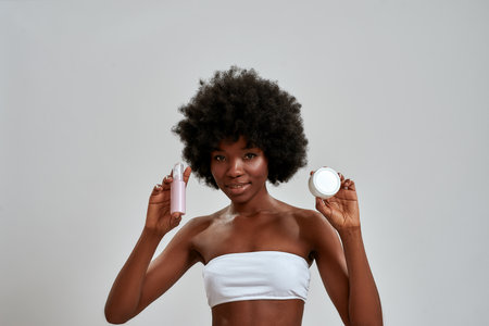 Portrait of cute young african american woman with afro hair smiling at camera, holding two skincare products while posing isolated over gray backgroundの写真素材