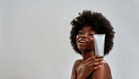Beautiful young half naked african american woman holding beauty product bottle on her shoulder, smiling at camera isolated over gray backgroundの写真素材