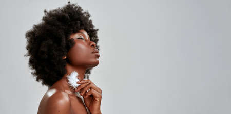 Portrait of sensual african american woman with afro hair touching her glowing skin with white feather, posing with eyes closed isolated over gray backgroundの写真素材