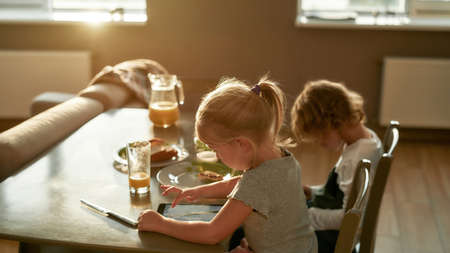 Cute little brother and sister using digital devices while having breakfast or lunch, sitting together at the table in the kitchenの写真素材