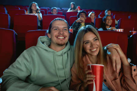 Portrait of cheerful young couple laughing, having drinks and popcorn while watching movie together, sitting in cinema auditoriumの写真素材
