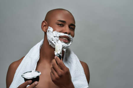 Shirtless attractive young african american man smiling at camera, using brush while applying shaving foam on his face, posing isolated over gray backgroundの写真素材