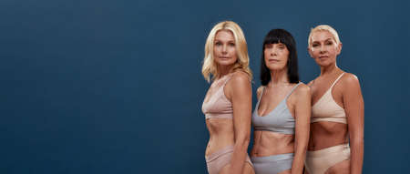 Feeling beautiful at any age. Three caucasian mature women in underwear looking at camera while posing half naked in studio against dark blue backgroundの写真素材
