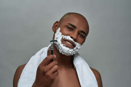 Handsome young african american man with shaving foam applied on his face smiling at camera, holding steel razor, posing isolated over gray backgroundの写真素材