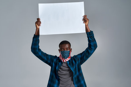 Young african american guy wearing bandana mask looking at camera, holding blank white banner ad above his head, asking for support, posing isolated over gray backgroundの写真素材