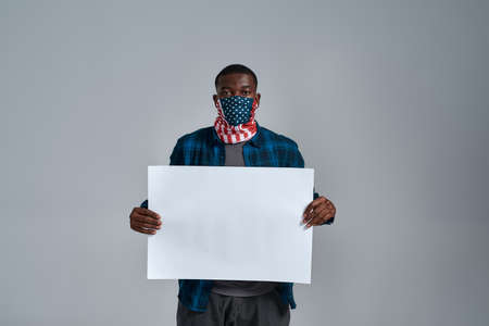 Young african american guy wearing bandana mask looking at camera, holding blank white banner ad in...