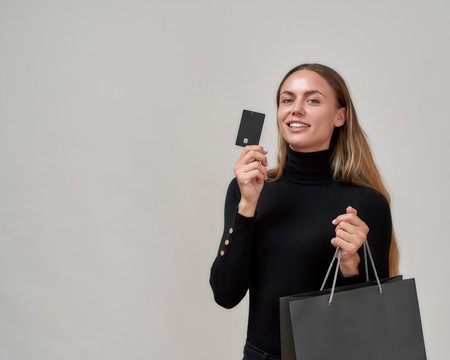 Gorgeous young woman wearing black clothes smiling at camera, holding plastic credit card while posing with shopping bag isolated over gray backgroundの写真素材