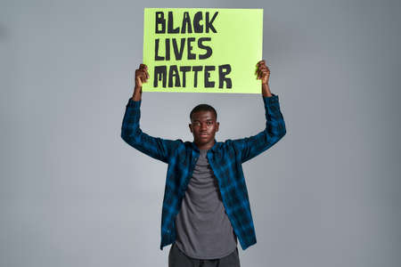 Confident young african american guy in casual clothes looking at camera, holding demonstration banner with BLM text above his head, posing isolated over gray backgroundの写真素材