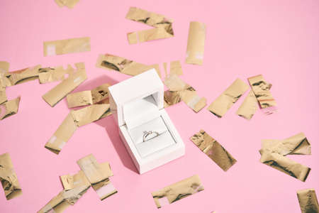 Wedding proposal. Elegant engagement diamond ring in white gift box and shiny golden confetti on pastel pink backgroundの写真素材