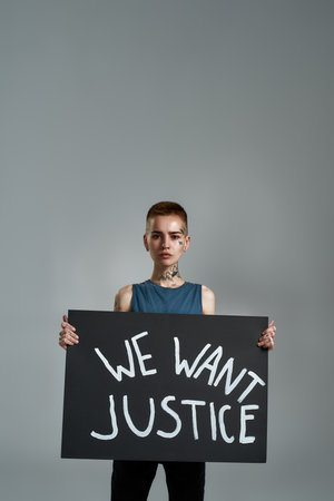 Confident tattooed young caucasian female activist looking at camera, holding cardboard banner with We want justice text, posing isolated over gray backgroundの写真素材