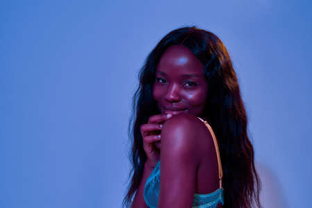 Portrait of cute african american young woman with long hair smiling at camera, looking shy while...