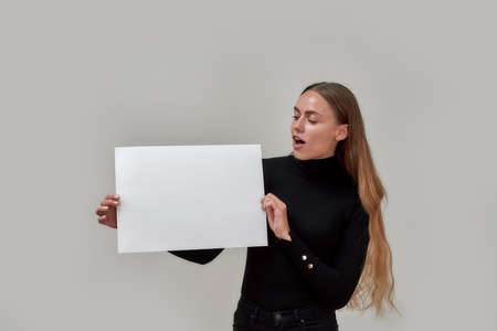 Excited young caucasian woman with long hair dressed in black displaying and looking at white blank...