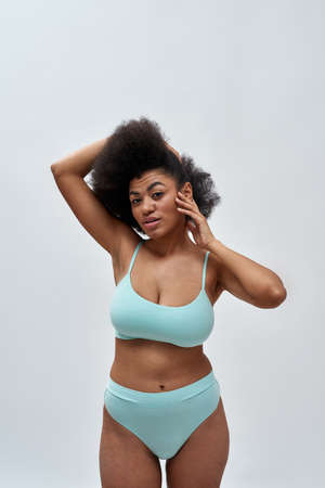 Curvy young mixed race female model with afro hair wearing blue underwear posing for camera, standing isolated over light backgroundの写真素材