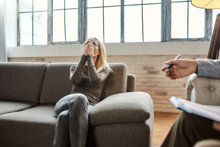 Depressed young caucasian woman on sofa beside psychotherapistの写真素材
