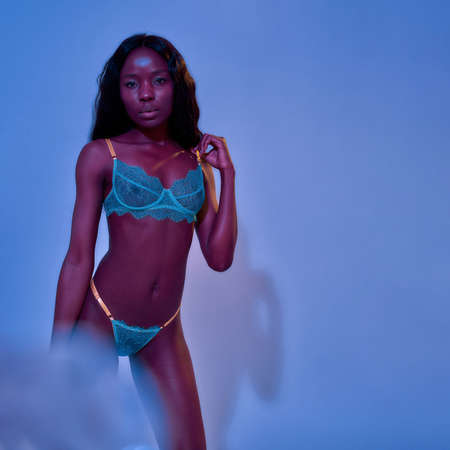 Hot african american young woman with perfect slim body wearing sexy lace lingerie looking at camera, adjusting bra strap while posing isolated in neon uv purple blue lightの写真素材