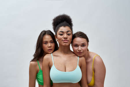 Portrait of three confident female friends, multiethnic young women in colorful underwear looking at camera, posing together isolated over light backgroundの写真素材