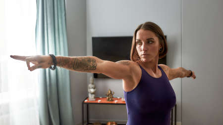 Portrait of sportive tattooed young woman in bodysuit looking focused while working out, doing...