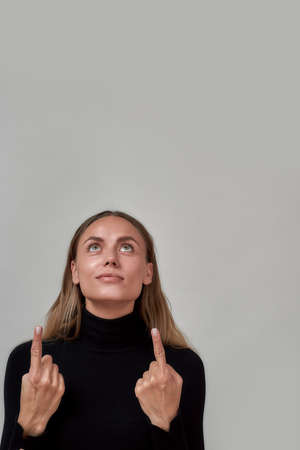 Portrait of pretty young caucasian woman dressed in black looking and pointing up at something, posing isolated over gray backgroundの写真素材