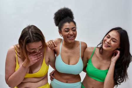 Three joyful diverse young women with different body shapes wearing colorful underwear laughing, posing together over light backgroundの写真素材
