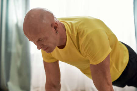 Online training. Mature man fitness instructor in sportswear standing in a plank while exercising at homeの写真素材